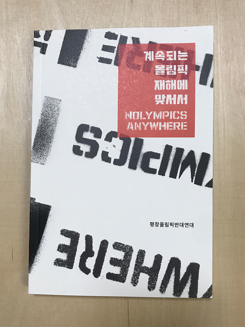 나무판 위에 책 한 권이 올려져 있다. 하얀색 표지에 비스듬히 검은색 영문 글자의 스텐실 이미지가 뒤집힌 모양으로 들어가 있고, 오른쪽 상단에 빨간색 네모칸 안에 흰색 글씨로 '계속되는 올림픽 재해에 맞서서 NOLYMPICS ANYWHERE'라고 적혀있다. 빨간 네모칸 아래쪽에는 작은 까만색 글씨로 '평창올림픽반대연대'라고 적혀있다. 